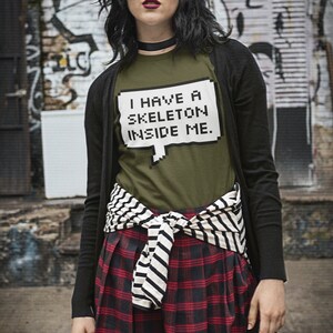May include: A woman wearing a black cardigan over a green t-shirt with the text "I have a skeleton inside me." in a pixelated font. She is wearing a red and black plaid skirt with a black and white striped belt tied around her waist.