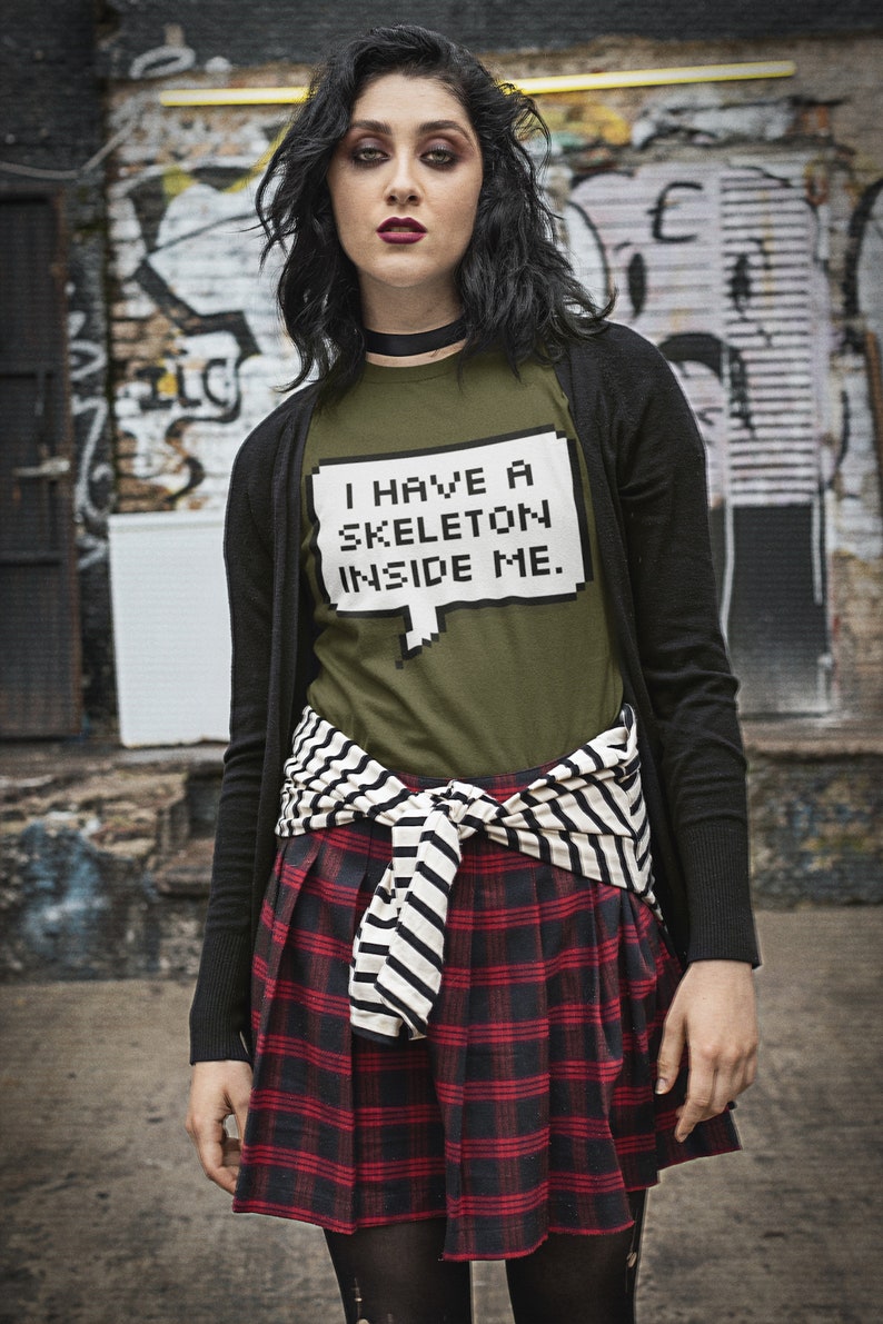 May include: A woman wearing a black cardigan over a green t-shirt with the text "I have a skeleton inside me." in a pixelated font. She is wearing a red and black plaid skirt with a black and white striped belt tied around her waist.