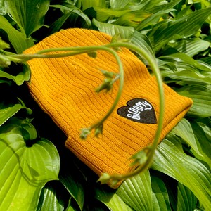 May include: A mustard yellow beanie with a black heart patch that says "BLOOMY" in white lettering.
