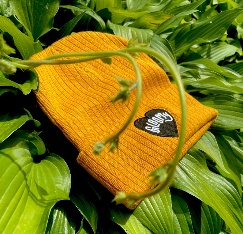 May include: A mustard yellow beanie with a black heart patch that says "BLOOMY" in white lettering.