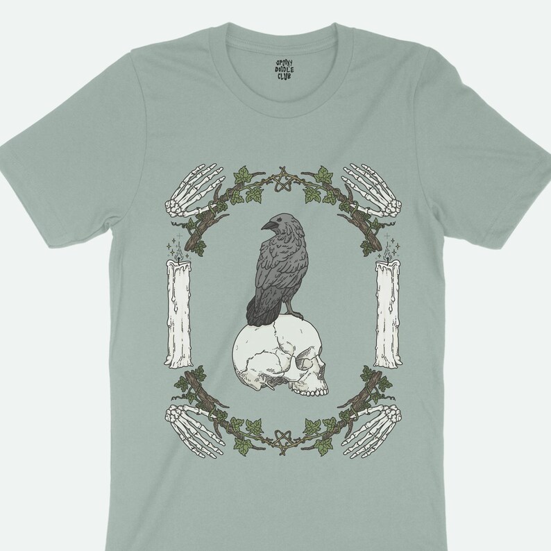 This dark academia shirt features a raven sitting on top of a skull. The macabre whimsigoth design is inspired by Edgar Allan Poe's The Raven.