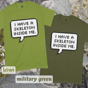 May include: Two t-shirts, one in kiwi green and one in military green, both featuring a pixelated speech bubble with the text "I have a skeleton inside me."