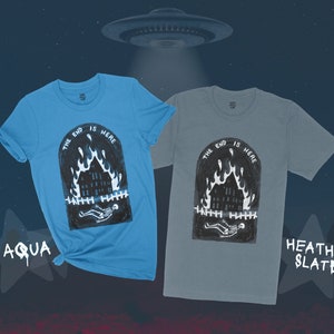 Op de afbeelding: Twee T-shirts met een grafische print van een brandend huis met een skelet dat ervoor ligt. De tekst "The end is here" is boven het huis gedrukt. De T-shirts zijn in de kleuren aqua en heather slate.