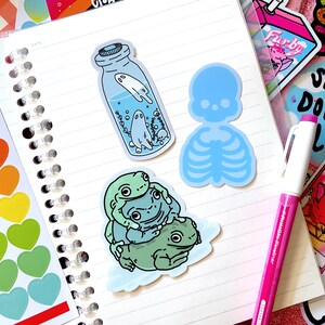 May include: Three frog stickers, a blue skeleton sticker, and a sticker of a glass jar with a ghost inside. The stickers are on a white notebook with a spiral binding.