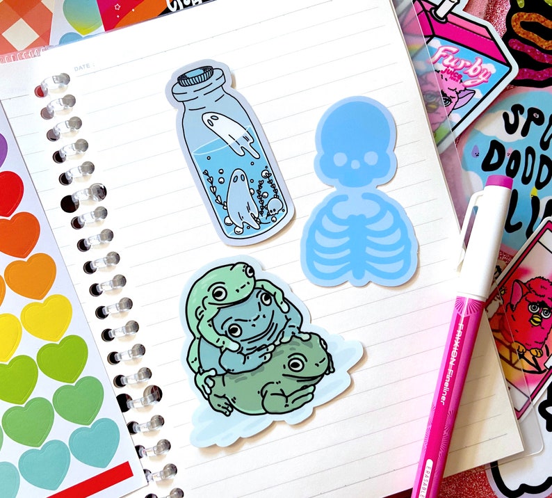 May include: Three frog stickers, a blue skeleton sticker, and a sticker of a glass jar with a ghost inside. The stickers are on a white notebook with a spiral binding.