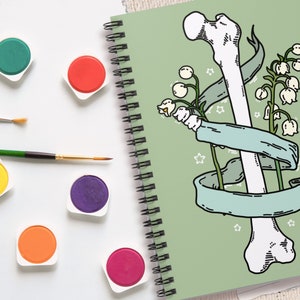 Op de afbeelding: Een groene spiraalnotitieblok met een wit bot en witte bloemen met groene stelen en bladeren. Het bot is omwikkeld met een lichtblauw lint. De achtergrond is lichtgroen.