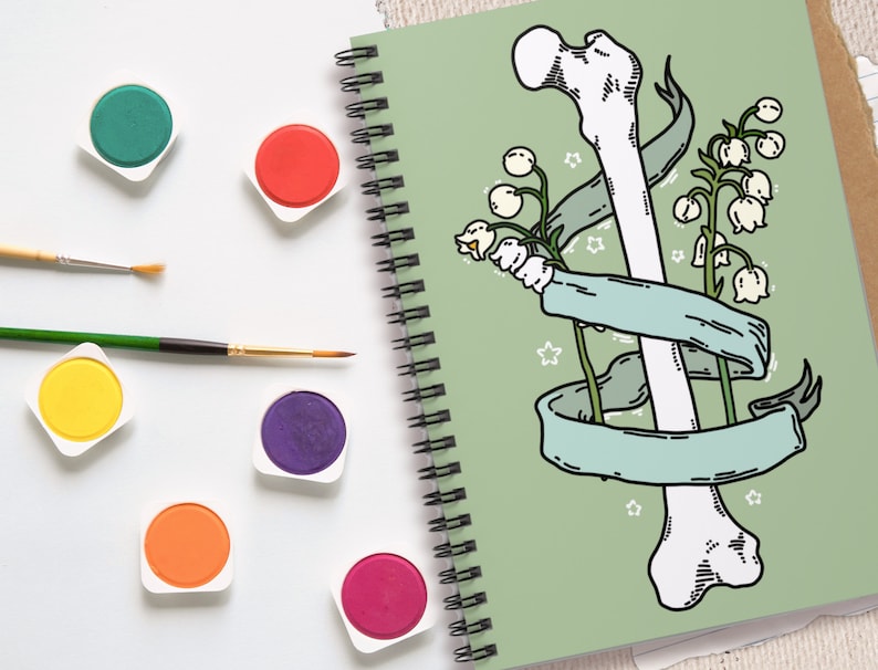 Op de afbeelding: Een groene spiraalnotitieblok met een wit bot en witte bloemen met groene stelen en bladeren. Het bot is omwikkeld met een lichtblauw lint. De achtergrond is lichtgroen.