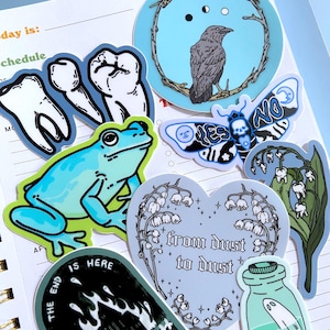 May include: A collection of stickers featuring various designs, including a frog, a crow, a moth, a heart with flowers, a bottle with a ghost, and a tombstone with flames. The stickers are on a blue and white notebook.
