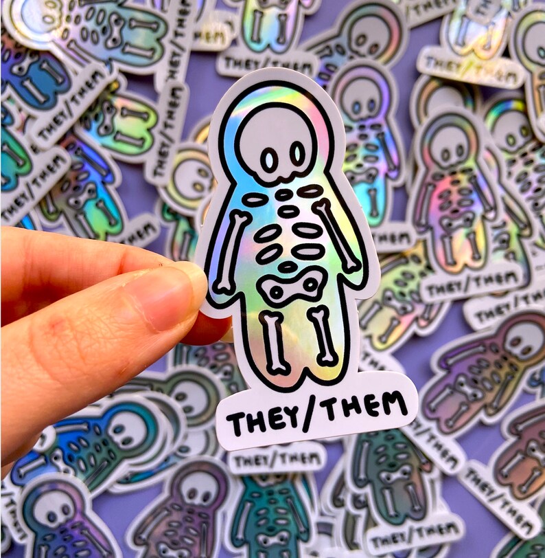 May include: A holographic sticker with a skeleton figure and the text "THEY/THEM". The sticker is surrounded by other similar stickers with different designs.