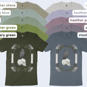May include: A collection of short-sleeved t-shirts in various colors, including military green and steel blue. Each shirt features a graphic of a raven perched on a skull, surrounded by a wreath and bone details.