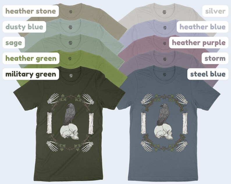 May include: A collection of short-sleeved t-shirts in various colors, including military green and steel blue. Each shirt features a graphic of a raven perched on a skull, surrounded by a wreath and bone details.