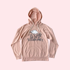 Pode incluir: Um moletom rosa claro com bolso canguru e capuz com cord&atilde;o. A frente apresenta um gr&aacute;fico branco de um gamb&aacute; e o texto "LIFE IS SCARY AND IT'S OK TO SCREAM ABOUT IT" em letras pretas.