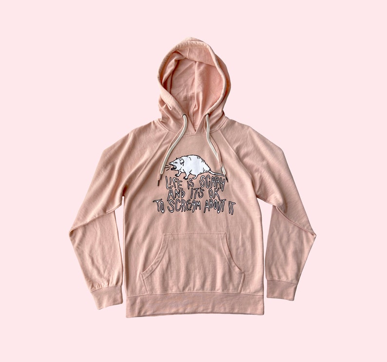 Pode incluir: Um moletom rosa claro com bolso canguru e capuz com cord&atilde;o. A frente apresenta um gr&aacute;fico branco de um gamb&aacute; e o texto "LIFE IS SCARY AND IT'S OK TO SCREAM ABOUT IT" em letras pretas.