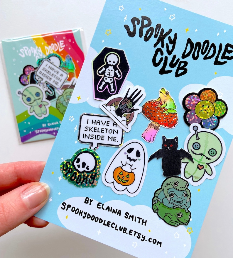 May include: A collection of spooky themed stickers featuring a variety of designs including a skeleton, a ghost, a bat, a frog, a flower, and a skull. The stickers are on a light blue background with the text "Spooky Doodle Club" in black.