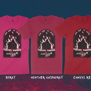 Op de afbeelding: Drie T-shirts met een grafische print van een brandend huis met een skelet dat er voor ligt. De tekst "The End is Here" is boven de print gedrukt. De T-shirts zijn in de kleuren bessen, heide-framboos en canvas rood.