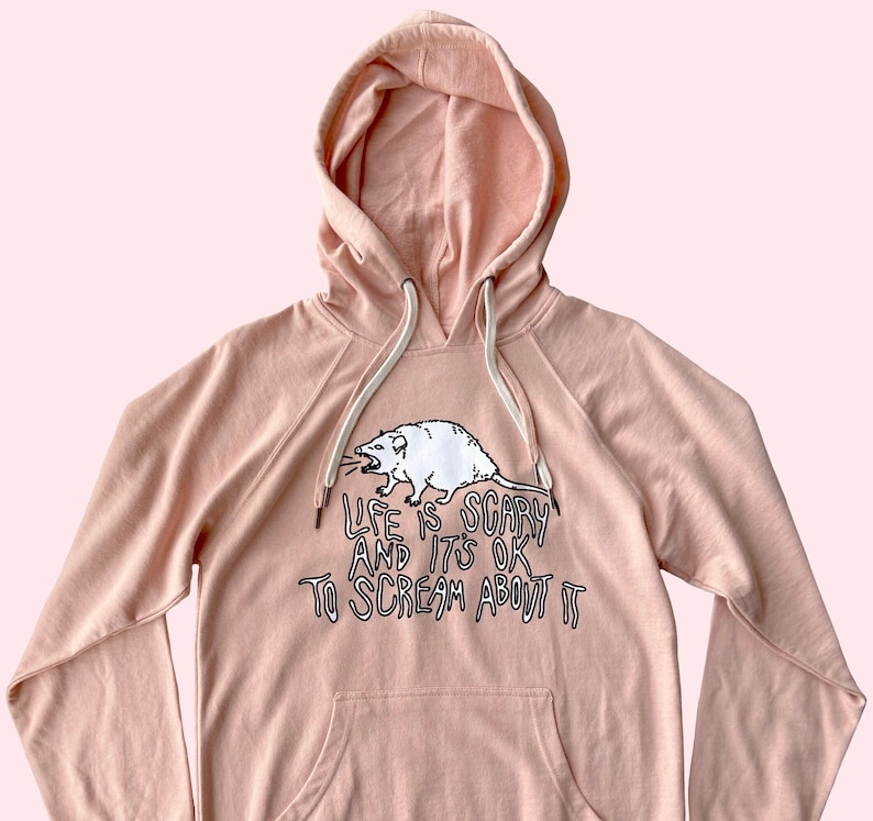 Pode incluir: Moletom rosa claro com bolso canguru e capuz com cord&atilde;o. A frente apresenta um gr&aacute;fico de gamb&aacute; branco e o texto "LIFE IS SCARY AND IT'S OK TO SCREAM ABOUT IT" em preto. O moletom &eacute; feito de material macio.