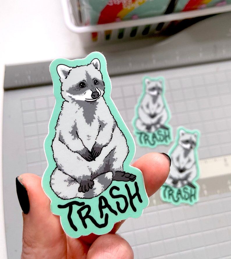 以下が含まれることがあります： アライグマのステッカーセット。メインのステッカーは、黒い「TRASH」の文字が入ったグレーと白のアライグマです。同じデザインの小さなステッカーが2つも見えます。ステッカーは薄い緑色の縁取りです。