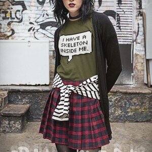 May include: A woman wearing a green t-shirt with the text "I have a skeleton inside me." in a pixelated speech bubble. She is also wearing a black cardigan, a black and white striped belt, a red and black plaid skirt, and black tights with holes in them. She is standing in front of a brick wall with graffiti.
