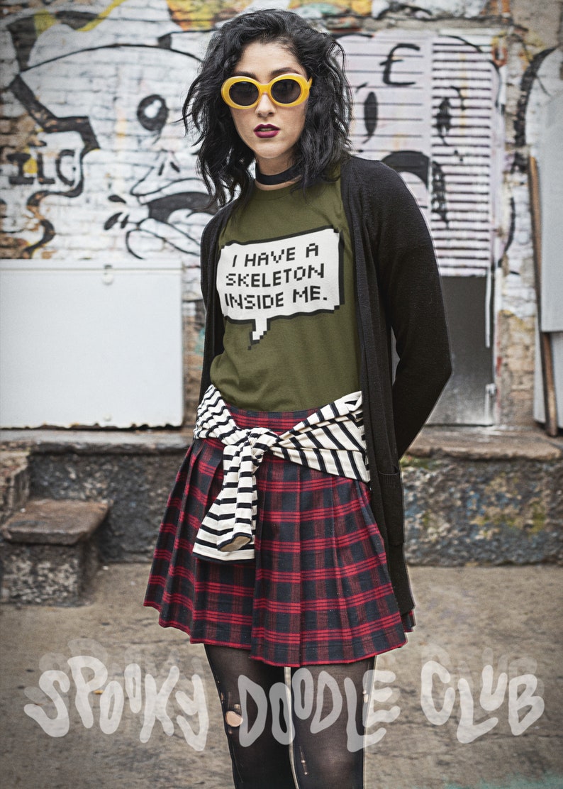 May include: A woman wearing a green t-shirt with the text "I have a skeleton inside me." in a pixelated speech bubble. She is also wearing a black cardigan, a black and white striped belt, a red and black plaid skirt, and black tights with holes in them. She is standing in front of a brick wall with graffiti.