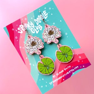 Op de afbeelding: Roze en groene oorhangers met een waterlelie- en waterleliebladontwerp. De oorbellen hebben een witte bloem met een gele kern en een groen waterlelieblad met waterdruppels. Op een roze en teal kaart met de tekst "Spooky Doodle Club".