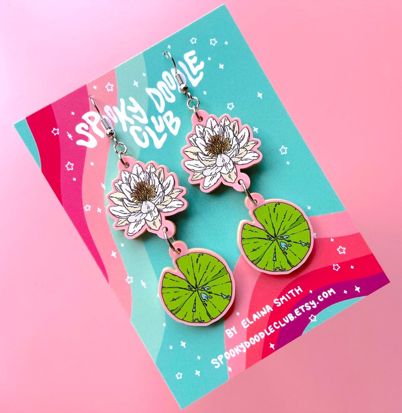 Op de afbeelding: Roze en groene oorhangers met een waterlelie- en waterleliebladontwerp. De oorbellen hebben een witte bloem met een gele kern en een groen waterlelieblad met waterdruppels. Op een roze en teal kaart met de tekst "Spooky Doodle Club".