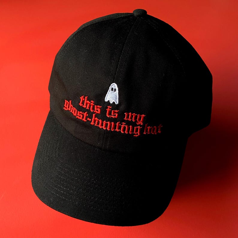 Op de afbeelding: Zwarte baseballpet met rode geborduurde tekst die "This is my ghost-hunting hat" zegt en een witte geestenafbeelding.