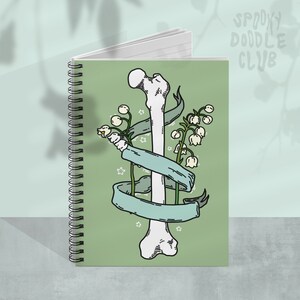 Op de afbeelding: Een groene spiraalnotitieblok met een ontwerp van een wit bot en een groen lint. Het bot is omwikkeld met een lint met witte bloemen. De notitieblokhoes heeft de tekst "Spooky Doodle Club" in wit.