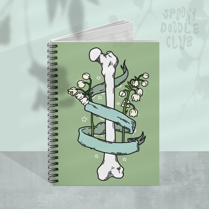 Op de afbeelding: Een groene spiraalnotitieblok met een ontwerp van een wit bot en een groen lint. Het bot is omwikkeld met een lint met witte bloemen. De notitieblokhoes heeft de tekst "Spooky Doodle Club" in wit.