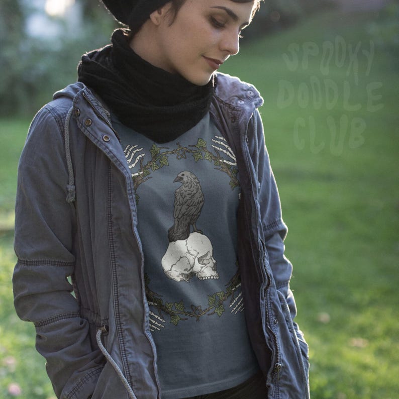 May include: A blue-grey t-shirt featuring a detailed illustration of a raven perched atop a skull, surrounded by a wreath of leaves and bones. The text "Spooky Doodle Club" is visible in the background. The shirt is part of a gothic or Halloween-themed collection.