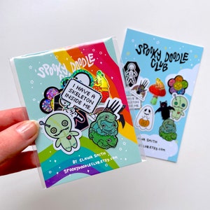 May include: A set of 10 enamel pins featuring spooky and cute designs. The pins include a skeleton, a ghost, a bat, a frog, a mushroom, a pumpkin, a flower, and a voodoo doll. The pins are packaged in a clear plastic bag with a rainbow background and the text "Spooky Doodle Club" and "By Elaina Smith".