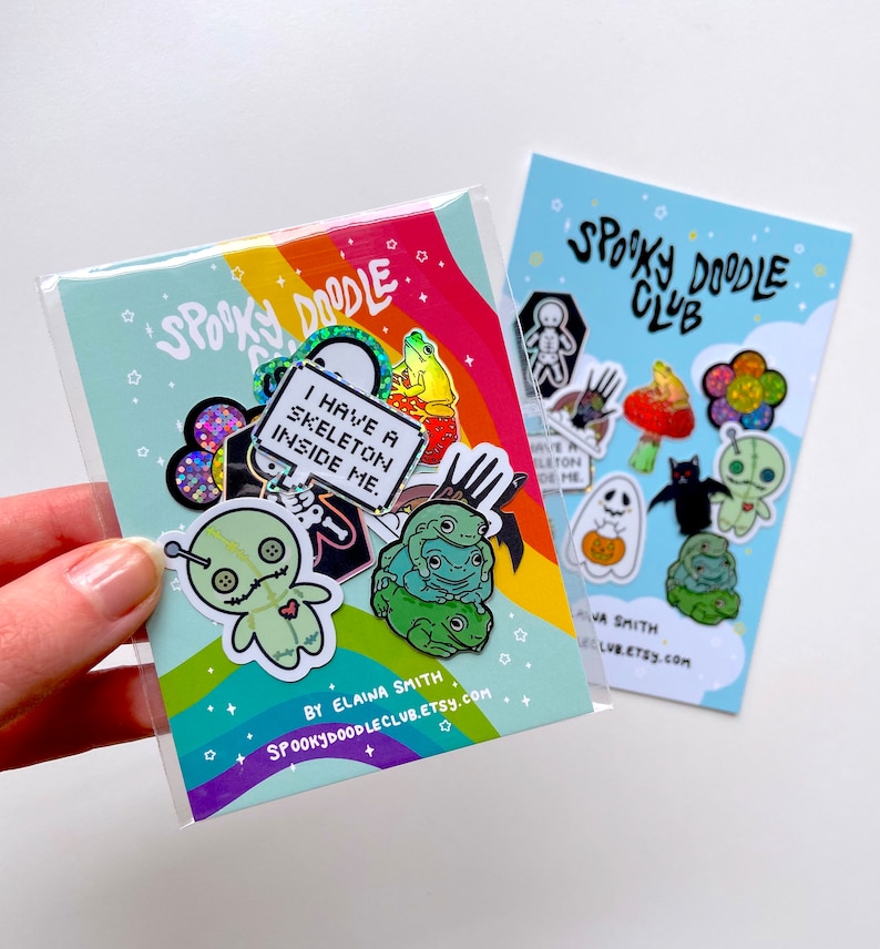 May include: A set of 10 enamel pins featuring spooky and cute designs. The pins include a skeleton, a ghost, a bat, a frog, a mushroom, a pumpkin, a flower, and a voodoo doll. The pins are packaged in a clear plastic bag with a rainbow background and the text "Spooky Doodle Club" and "By Elaina Smith".