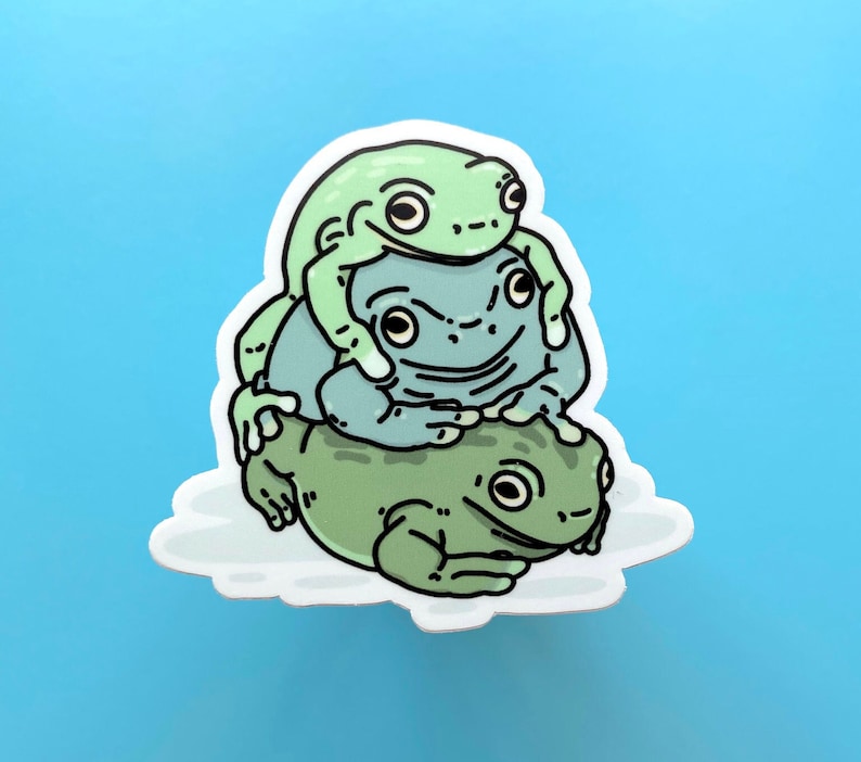 May include: A sticker featuring three green frogs stacked on top of each other. The top frog is green with black eyes and a wide smile. The middle frog is blue with black eyes and a closed mouth. The bottom frog is green with black eyes and a closed mouth.