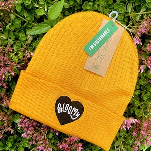 May include: A mustard yellow beanie with a black heart-shaped patch that says "GLOOMY" in white lettering. The beanie has a tag attached that says "I'm Sustainable" and "Recycled Polyester".