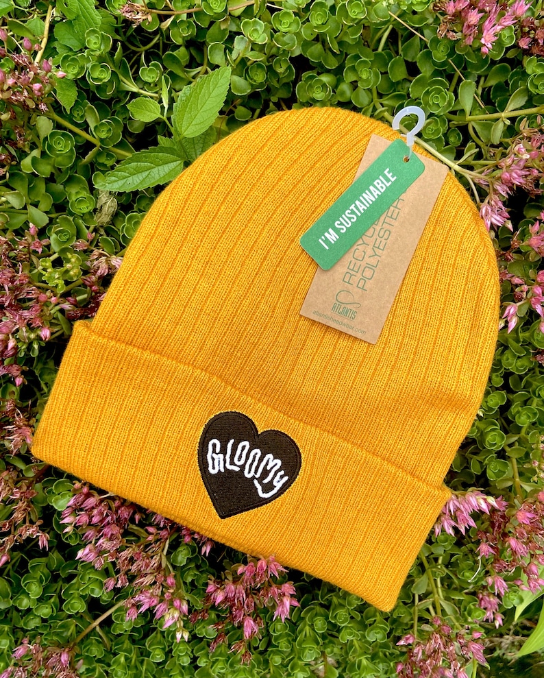 May include: A mustard yellow beanie with a black heart-shaped patch that says "GLOOMY" in white lettering. The beanie has a tag attached that says "I'm Sustainable" and "Recycled Polyester".