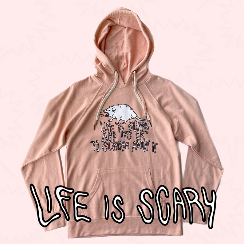 Pode incluir: Um moletom rosa claro com cord&atilde;o branco e bolso canguru. O texto "LIFE IS SCARY" &eacute; impresso em negrito e com contorno. Acima do bolso, um desenho de um gamb&aacute; branco e o texto "LIFE IS SCARY AND IT'S OK TO SCREAM ABOUT IT."
