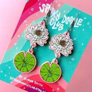 Op de afbeelding: Een paar bungelende oorbellen met een wit en geel waterlelie-ontwerp en een groen waterlelieblad. De oorbellen zijn roze met zilveren haken. De achtergrond is roze en regenboogkleurig met de tekst "Spooky Doodle Club".