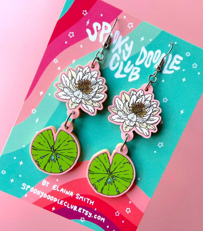Op de afbeelding: Een paar bungelende oorbellen met een wit en geel waterlelie-ontwerp en een groen waterlelieblad. De oorbellen zijn roze met zilveren haken. De achtergrond is roze en regenboogkleurig met de tekst "Spooky Doodle Club".