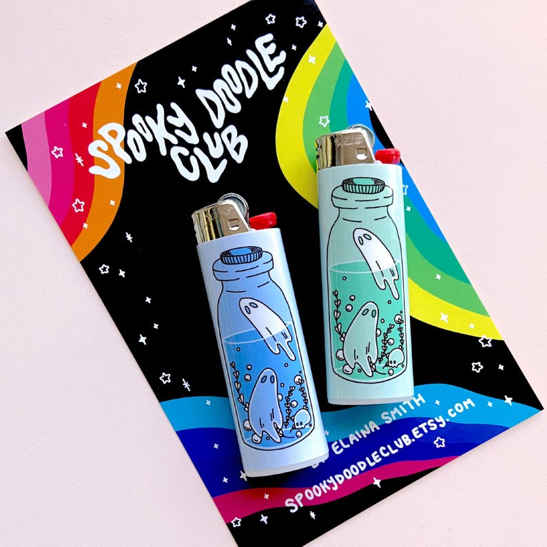 Puede incluir: Dos encendedores azules y verdes con ilustraciones de fantasmas dentro de botellas de vidrio. Los encendedores est&aacute;n sobre un fondo negro con rayas de arco&iacute;ris y estrellas. El texto "Spooky Doodle Club" est&aacute; escrito en blanco sobre el fondo.