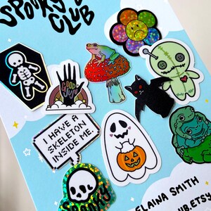 May include: A set of 10 holographic stickers featuring spooky and cute designs. The stickers include a skeleton in a coffin, a frog on a mushroom, a ghost holding a pumpkin, a voodoo doll, a bat, a hand with the words "Good Juju", a skull with the word "Spooky", a flower with a sad face, and two frogs hugging. The stickers are on a blue background with white stars.