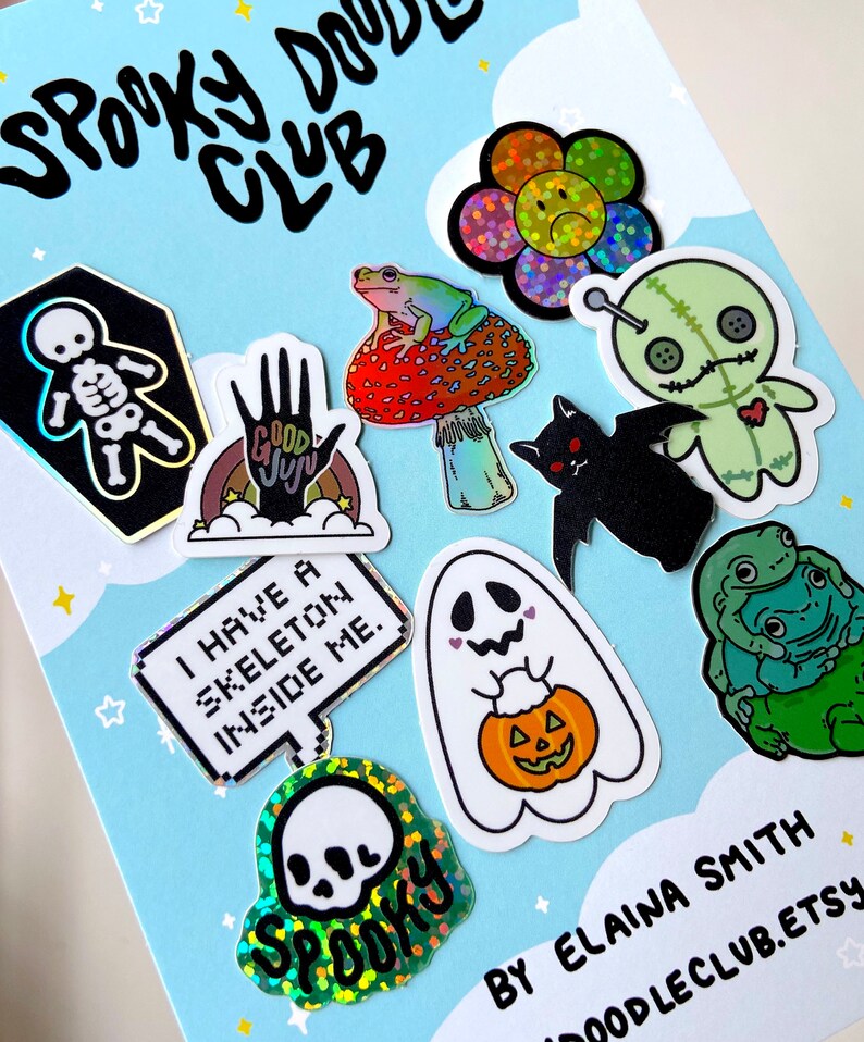 May include: A set of 10 holographic stickers featuring spooky and cute designs. The stickers include a skeleton in a coffin, a frog on a mushroom, a ghost holding a pumpkin, a voodoo doll, a bat, a hand with the words "Good Juju", a skull with the word "Spooky", a flower with a sad face, and two frogs hugging. The stickers are on a blue background with white stars.
