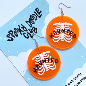 Może przedstawiać: Okrągłe kolczyki w kolorze pomarańczowym z białym szkieletem i napisem "HAUNTED" w kolorze czarnym. Kolczyki znajdują się na niebieskim tle z białymi gwiazdkami.