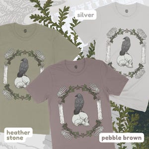May include: Three t-shirts in heather stone, pebble brown, and silver. Each shirt features a graphic of a raven perched on a skull, surrounded by a wreath of leaves, candles, and skeletal hands. The shirts are short-sleeved.