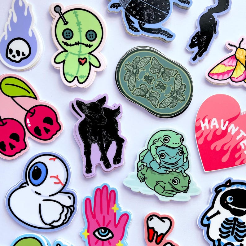May include: A collection of colorful stickers featuring various designs, including a voodoo doll, a goat, a skull, a heart with the word "HAUNTED" on it, a hand with an eye, a group of frogs, and a cherry with a skull on it.