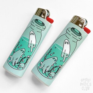 Puede incluir: Dos encendedores turquesa con un dise&ntilde;o de una botella de vidrio llena de un l&iacute;quido verde y fantasmas blancos. El texto "Spooky Doodle Club" est&aacute; impreso en la parte inferior de un encendedor.