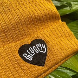 May include: A mustard yellow beanie with a black heart-shaped patch that says "BLOOMY" in white embroidery.