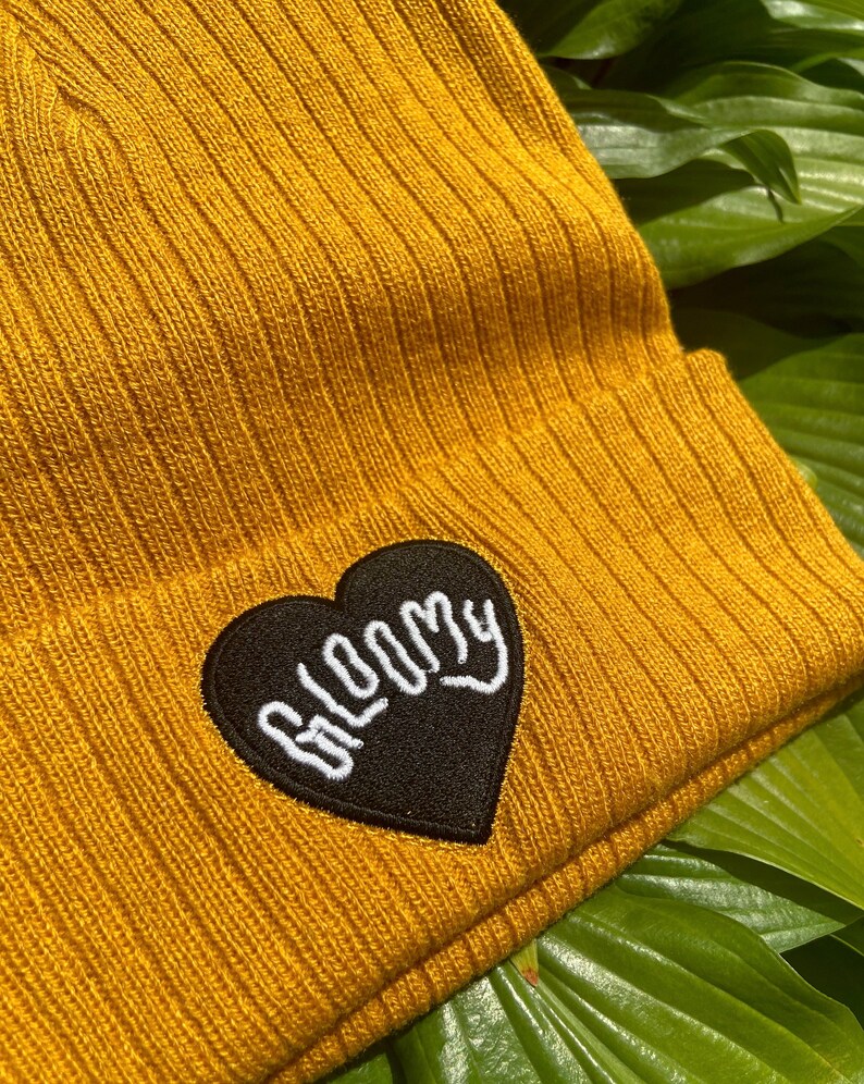May include: A mustard yellow beanie with a black heart-shaped patch that says "BLOOMY" in white embroidery.