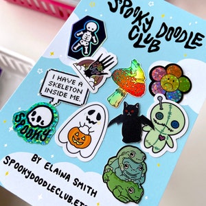 May include: A card with the text "Spooky Doodle Club" and a variety of holographic stickers featuring spooky and cute designs, including a skeleton, a ghost, a pumpkin, a frog, a flower, a bat, and a voodoo doll. The card also includes the text "By Elaina Smith" and "Spookydoodleclub.etsy.com".