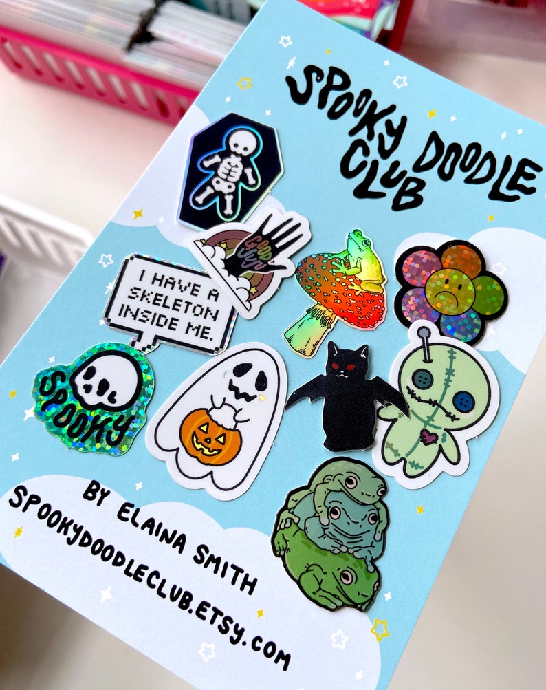 May include: A card with the text "Spooky Doodle Club" and a variety of holographic stickers featuring spooky and cute designs, including a skeleton, a ghost, a pumpkin, a frog, a flower, a bat, and a voodoo doll. The card also includes the text "By Elaina Smith" and "Spookydoodleclub.etsy.com".