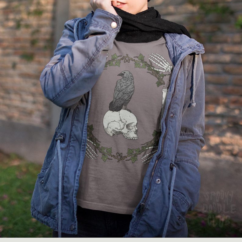 May include: A gray t-shirt with a black raven perched on a skull. The raven and skull are surrounded by a wreath of green leaves and skeletal hands. The text 'Spooky Doodle Crew' is visible in the bottom right corner of the image.