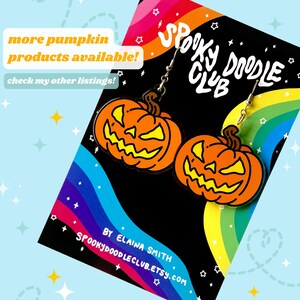 May include: A pair of pumpkin-shaped earrings with a spooky doodle club logo on the card. The earrings are orange with a yellow face and black eyes. The card is rainbow colored with white stars.